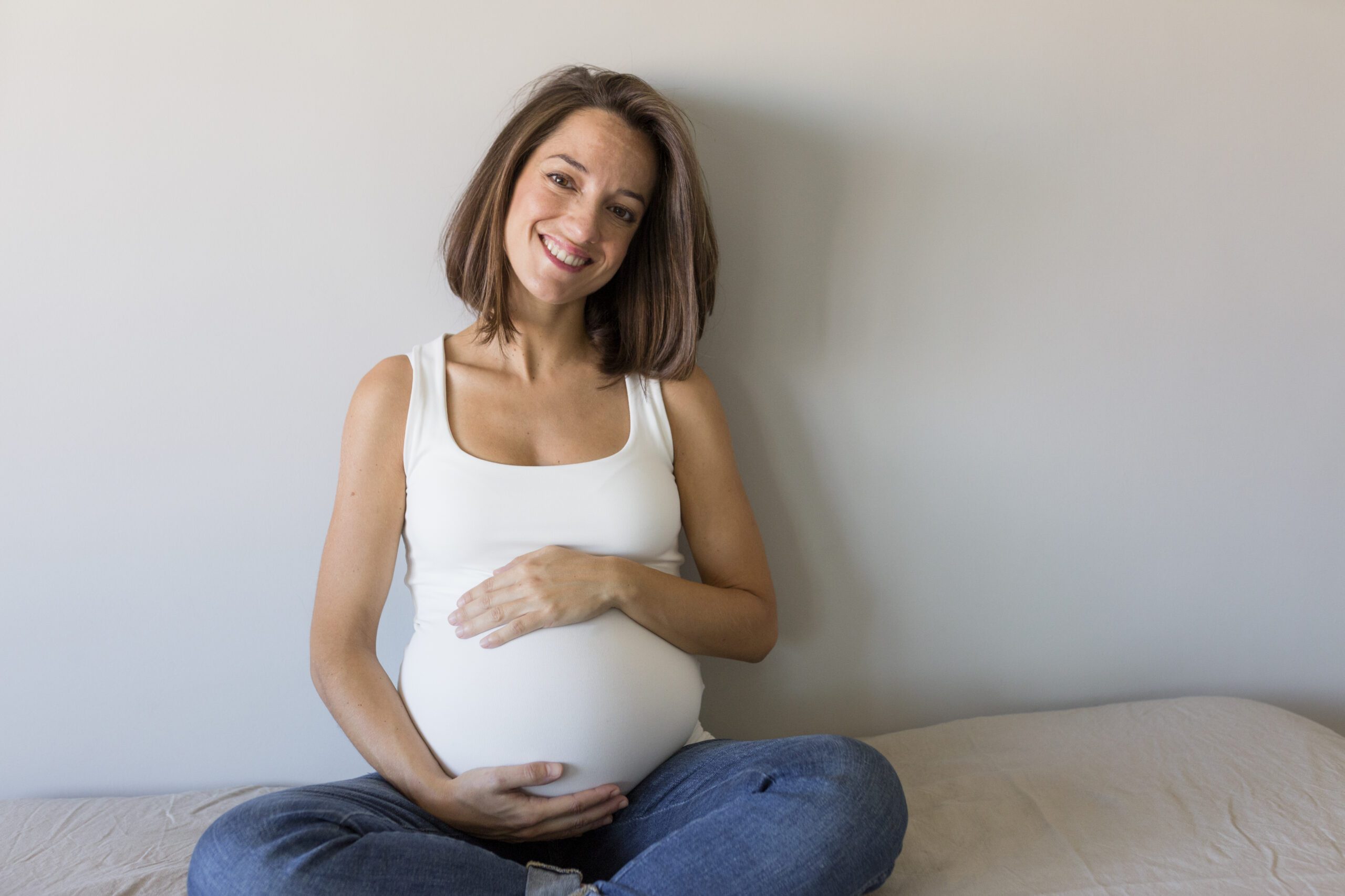 Attractive pregnant woman is sitting in bed, holding her belly and smiling. Last months of pregnancy.Lifestyle indoors.